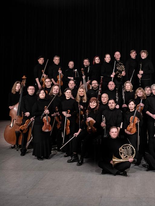 Das Orchester steht und sitzt in einer wolkenartigen Formation beieinander. Alle haben ihre Instrumente dabei.