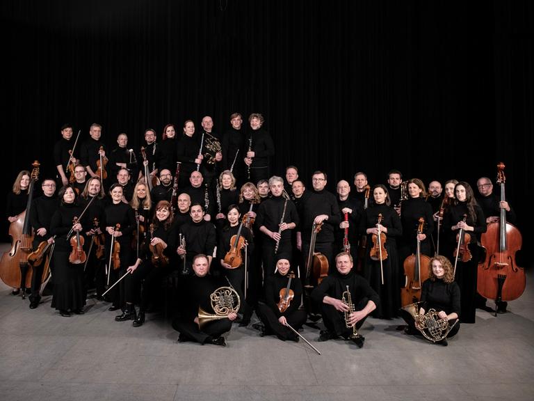 Das Orchester steht und sitzt in einer wolkenartigen Formation beieinander. Alle haben ihre Instrumente dabei.