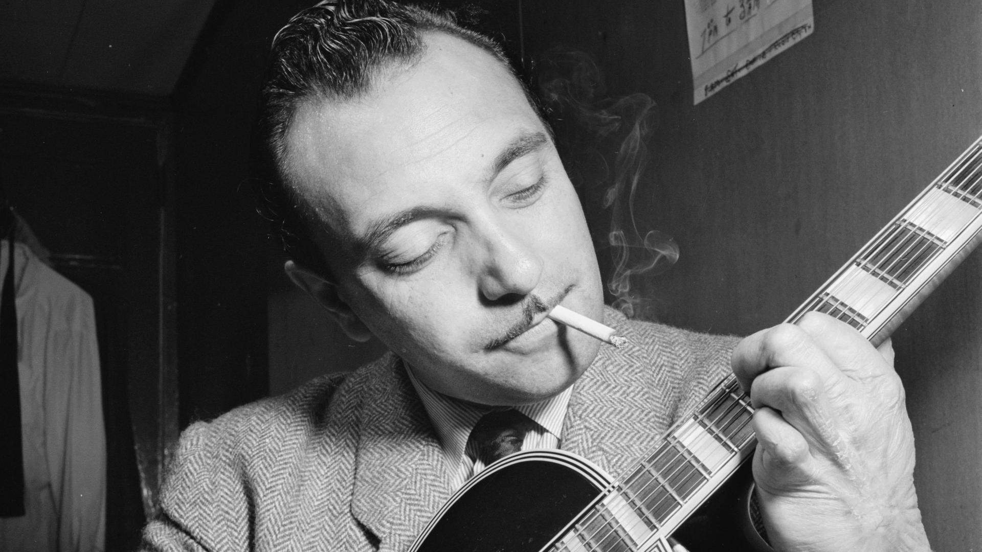 Ein schwarzweißes Portrait des Jazz-Musikers Django Reinhardt in einem New Yorker Jazzclub. Es zeigt seinen Oberkörper und Kopf, seine Gitarre ist im Vordergrund. Er spielt und schaut mit einem sanften Lächeln zu den Saiten. Im Mund hat er eine Zigarette.