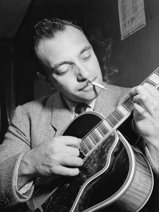 Ein schwarzweißes Portrait des Jazz-Musikers Django Reinhardt in einem New Yorker Jazzclub. Es zeigt seinen Oberkörper und Kopf, seine Gitarre ist im Vordergrund. Er spielt und schaut mit einem sanften Lächeln zu den Saiten. Im Mund hat er eine Zigarette.