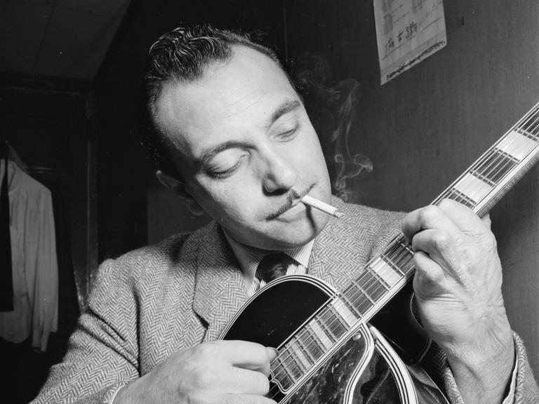 Ein schwarzweißes Portrait des Jazz-Musikers Django Reinhardt in einem New Yorker Jazzclub. Es zeigt seinen Oberkörper und Kopf, seine Gitarre ist im Vordergrund. Er spielt und schaut mit einem sanften Lächeln zu den Saiten. Im Mund hat er eine Zigarette.