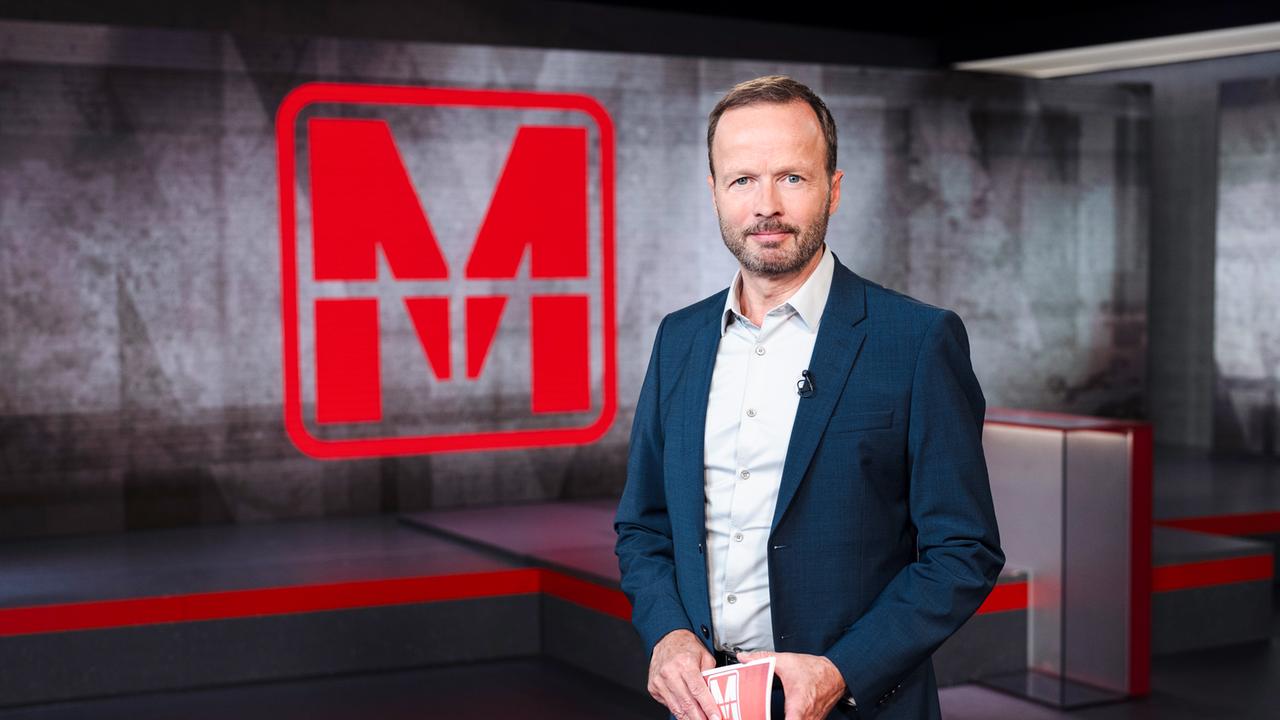 WDR-Journalist Georg Restle steht mit Moderationskarte in der Hand vor einer Wand, auf der das Monitor-Logo zu sehen ist.