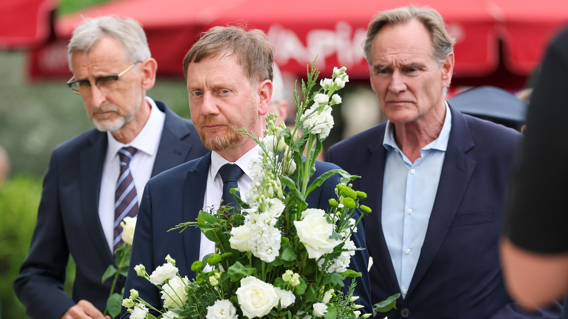 Drei Männer bei einem Gedenken mit Blumen.