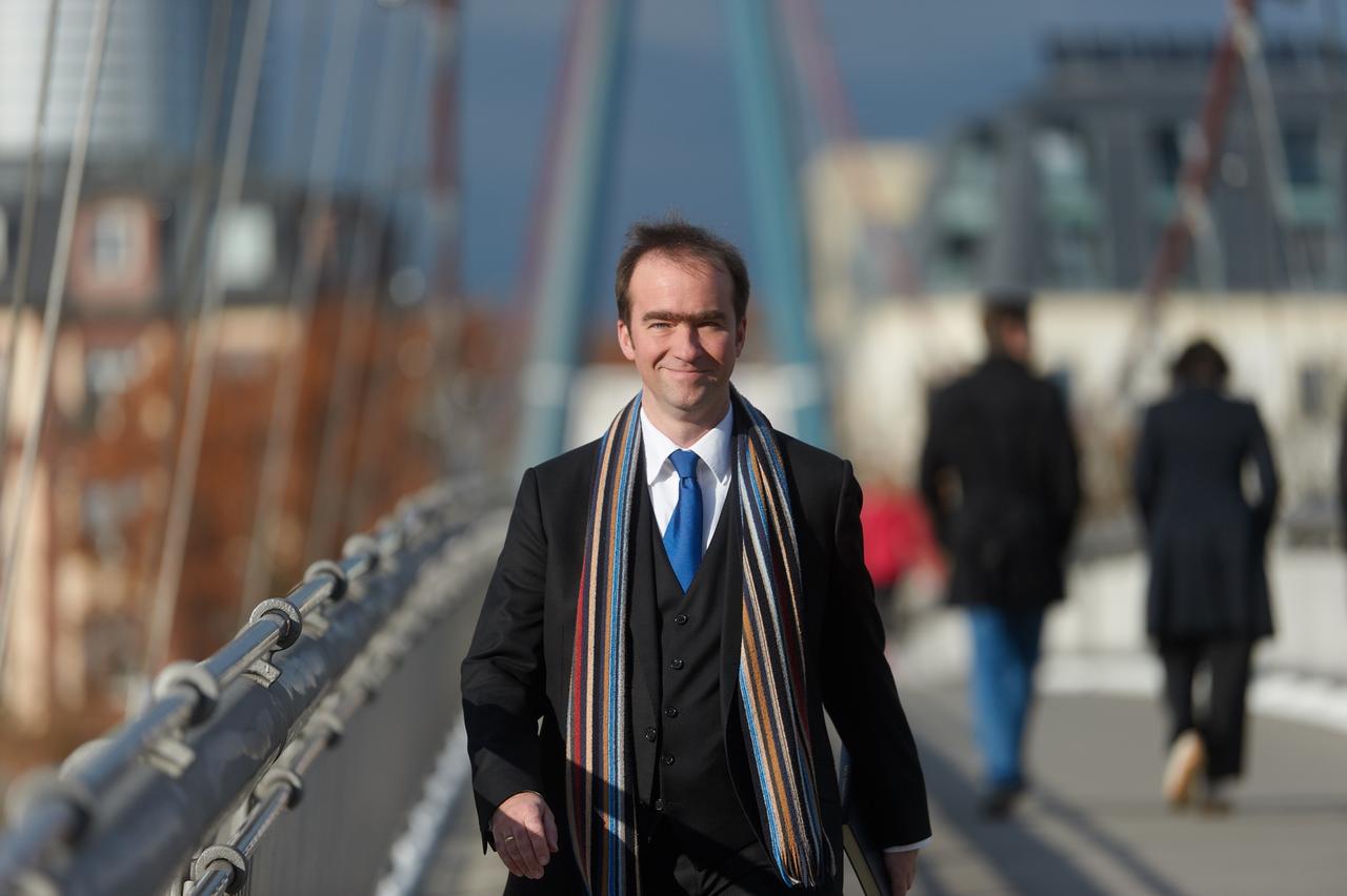 Georg Poplutz spaziert über eine der Hambuger Brücken und lacht dabei im Mantel und mit buntem Schal in die Kamera.