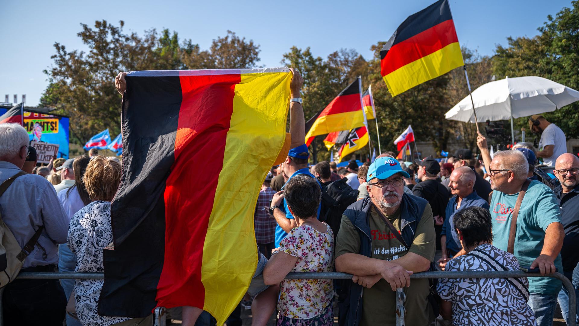 Viele ältere Menschen stehen mit Deutschlandfahnen und AfD-Schildern auf einem Platz. Unmittelbar im Vordergrund befindet sich ein Absperrgitter. Drei Frauen mit wild gemusterten Kleidern sind ebenfalls sichtbar.