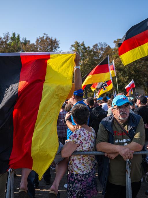 Viele ältere Menschen stehen mit Deutschlandfahnen und AfD-Schildern auf einem Platz. Unmittelbar im Vordergrund befindet sich ein Absperrgitter. Drei Frauen mit wild gemusterten Kleidern sind ebenfalls sichtbar.