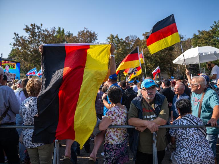 Viele ältere Menschen stehen mit Deutschlandfahnen und AfD-Schildern auf einem Platz. Unmittelbar im Vordergrund befindet sich ein Absperrgitter. Drei Frauen mit wild gemusterten Kleidern sind ebenfalls sichtbar.