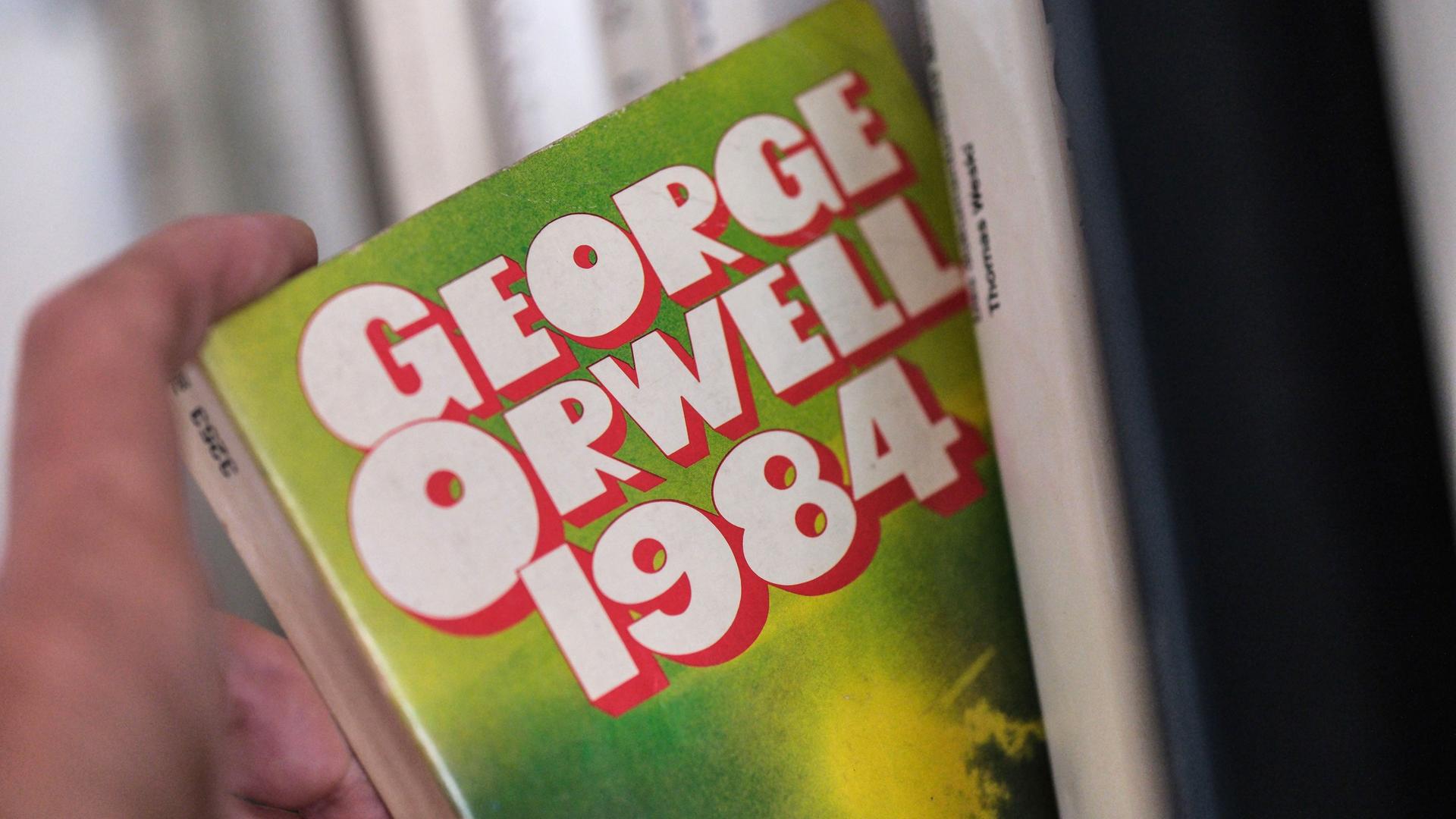 Ein ältere Ausgabe des Romans "1984" von George Orwell wird von einer Hand aus einem Bücherregal genommen. 