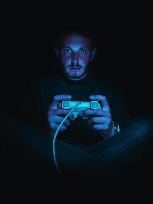 Ein Mann sitzt in einer dunklen Umgebung und hält einen Gaming-Controller. Seine Gesichtshaut ist rötlich, seine Augen sind weit aufgerissen.
