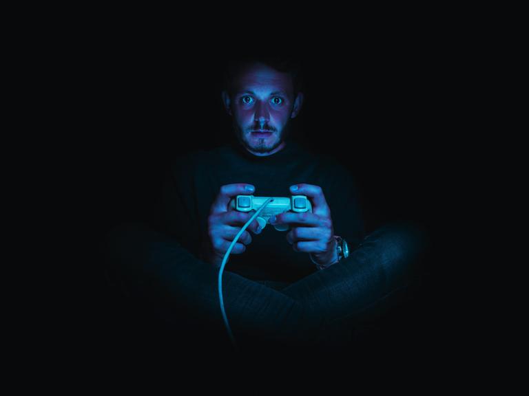 Ein Mann sitzt in einer dunklen Umgebung und hält einen Gaming-Controller. Seine Gesichtshaut ist rötlich, seine Augen sind weit aufgerissen.