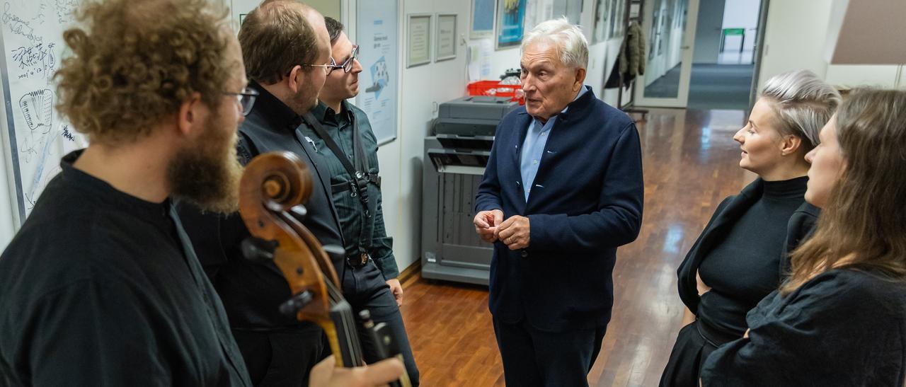 Ein älterer Mann steht auf einem Büroflur, von Musikern umringt, die mit ihm ins Gespräch vertieft sind.
