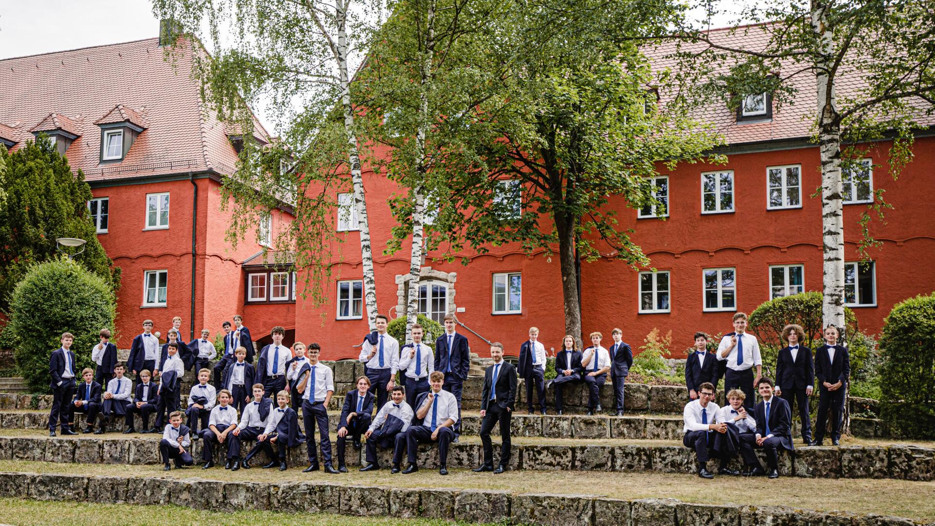 Eine große Gruppe Jungs steht auf dem Rasen einer weitläufigen Anlage, die von roten Gebäuden dominiert wird.