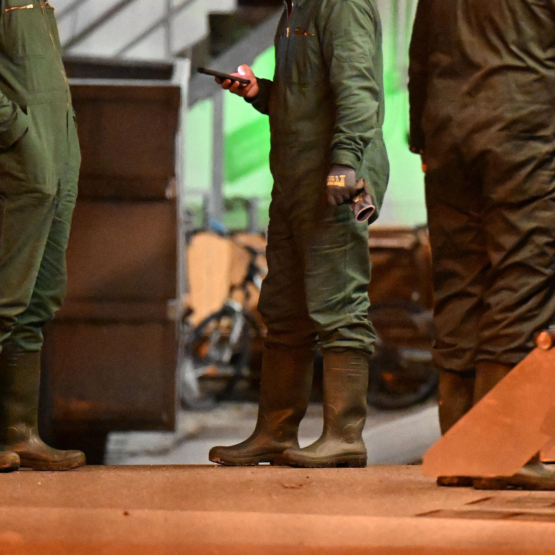 Man sieht drei Personen in Gummistiefeln und grünen Schutzanzügen im Gespräch in einem landwirtschaftlichen Gebäude. Ihre Köpfe sind abgeschnitten. Man sieht drei Personen in Gummistiefeln und grünen Schutzanzügen im Gespräch in einem landwirtschaftlichen Gebäude. Ihre Köpfe sind abgeschnitten.