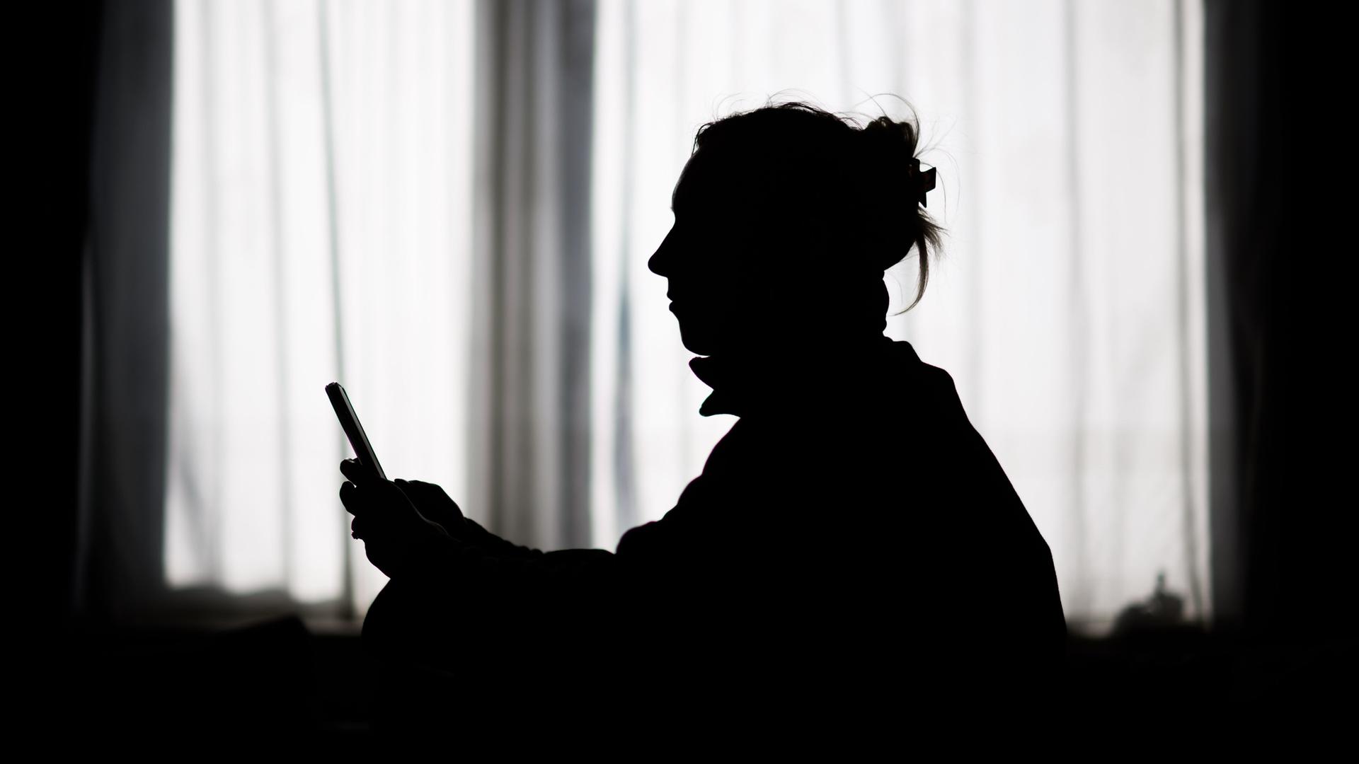 Die Silhouette einer Frau ist vor einem Fenster mit geschlossenen Vorhängen zu erkennen. Sie schaut auf ein Smartphone, das sie in der Hand hält.
