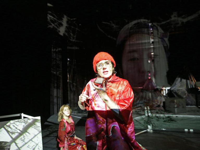 Szenenbild aus der Aufführung von "Mauern" des Künstler*innenkollektivs "She She Pop". Zu sehen sind verschiedene Überblendungen. Im Vordergrund eine Person mit roter Mütze, die einen Spiegel hält. Links davon, knieend, eine Frau im roten Kleid. Rechts im Hintergrund ein eingeblendetes Gesicht. 