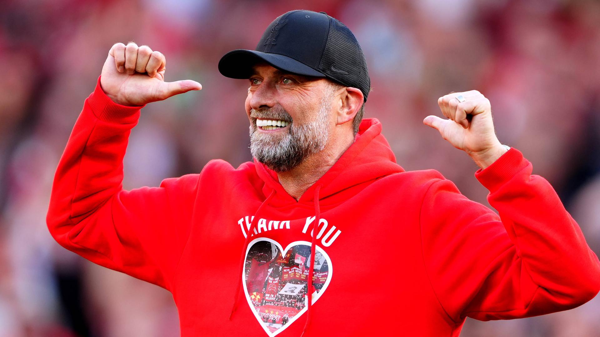 Jürgen Klopp trägt einen roten Kapuzen-Pullover und ein schwarzes Cap und zeigt mit den Daumen auf sich.