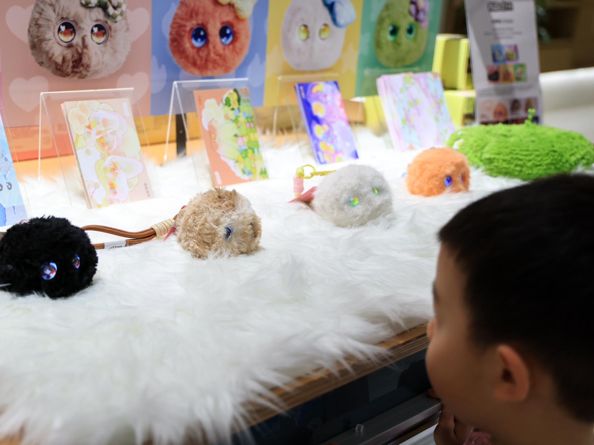 Ein chinesisches Kind schaut auf mehrere KI-Spielzeuge der Marke „Fuzozo“. Das sind kleine, plüschige KI-Begatl-Roboter, die zu sprachbasierter Interaktion fähig sind und mit Berührungssensoren ausgestattet. Ein chinesisches Kind schaut auf mehrere KI-Spielzeuge der Marke „Fuzozo“. Das sind kleine, plüschige KI-Begatl-Roboter, die zu sprachbasierter Interaktion fähig sind und mit Berührungssensoren ausgestattet.