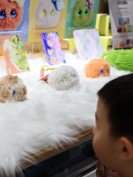 Ein chinesisches Kind schaut auf mehrere KI-Spielzeuge der Marke „Fuzozo“. Das sind kleine, plüschige KI-Begatl-Roboter, die zu sprachbasierter Interaktion fähig sind und mit Berührungssensoren ausgestattet. 
