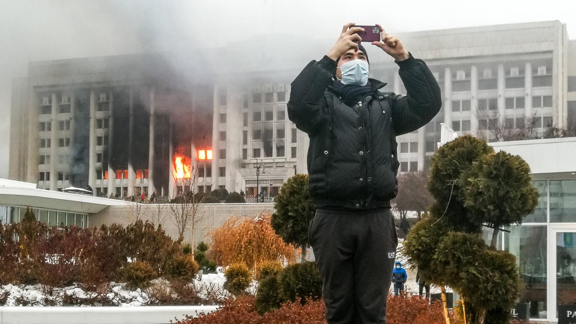 Ein junger Mann macht in der kasachischen Stadt Almaty mit seinem Smartphone ein Foto, im Hintergrund brennt das Amtsgebäude des Bürgermeisters
