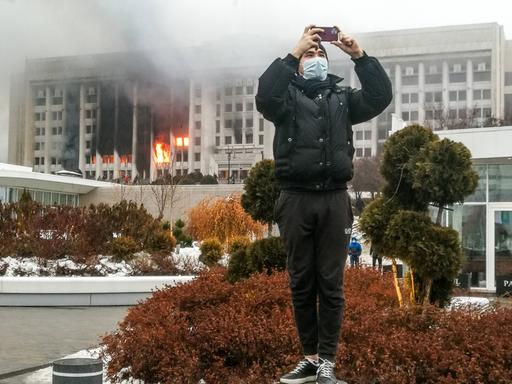 Ein junger Mann macht in der kasachischen Stadt Almaty mit seinem Smartphone ein Foto, im Hintergrund brennt das Amtsgebäude des Bürgermeisters