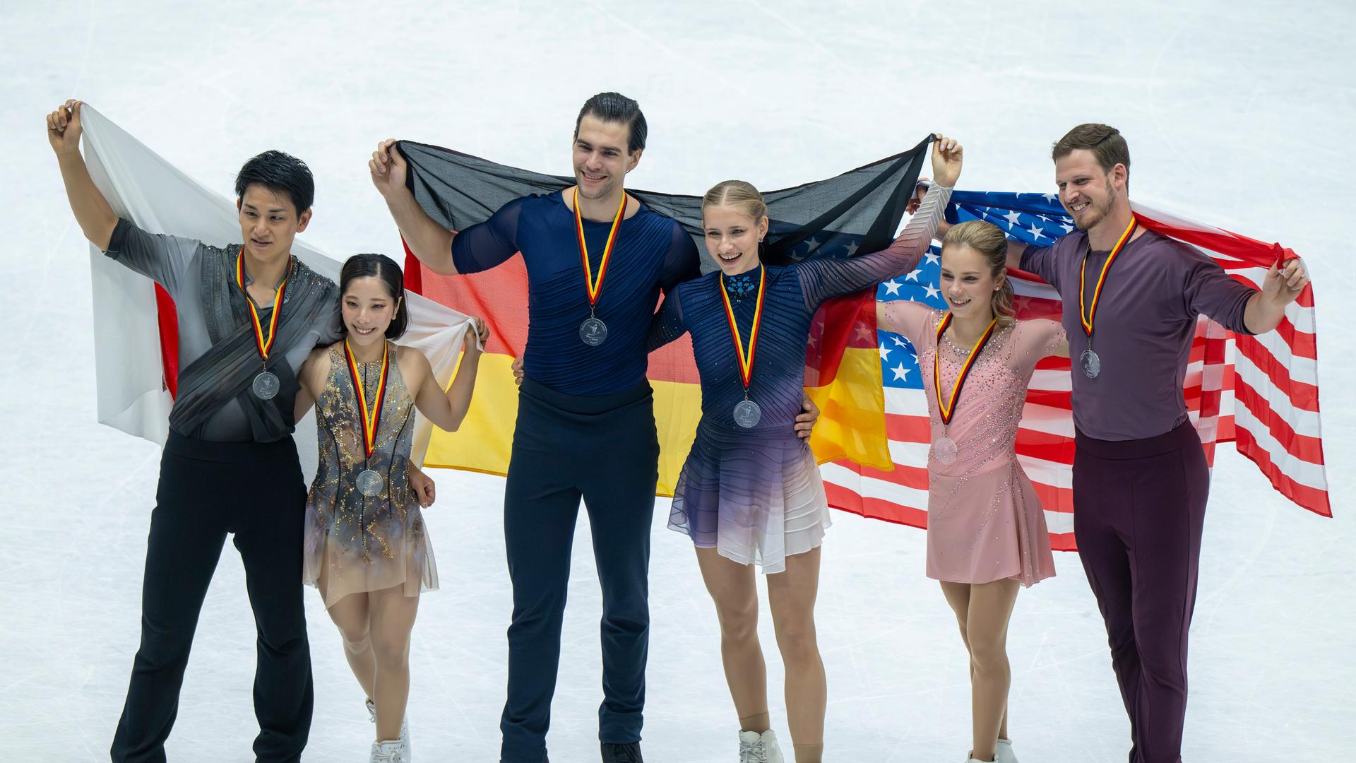 Minerva-Fabienne Hase und Nikita Volodin stehen als Sieger auf dem Podest. Neben ihnen stehen die Japaner Riku Miura und Ryuichi Kihara sowie Alisa Efimova und Misha Mitrofanov aus USA. Die Paare halten die Landesflaggen in die Höhe.
