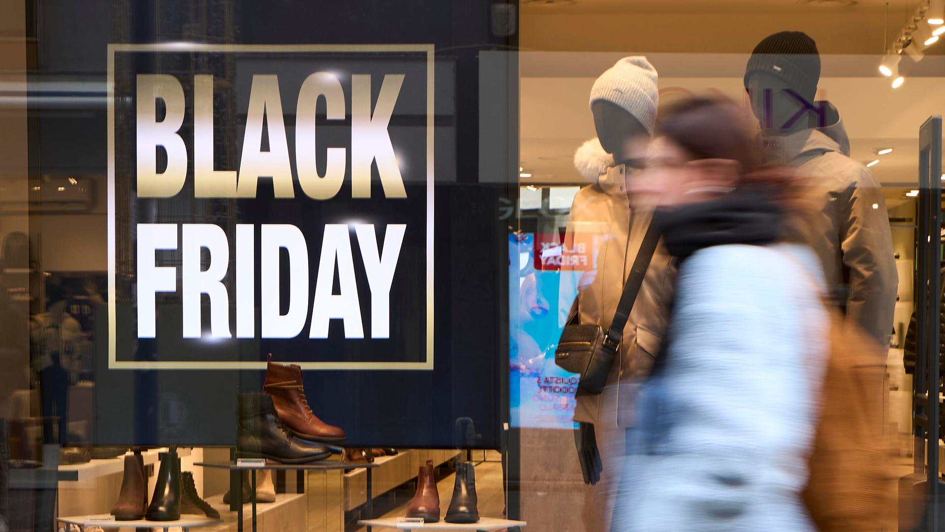 Fußgänger in Winterkleidung laufen an einem hell erleuchteten Schuhladen vorbei; im Schaufenster sind ein großes „BLACK FRIDAY“-Schild und Lederschuhe zu sehen. Fußgänger in Winterkleidung laufen an einem hell erleuchteten Schuhladen vorbei; im Schaufenster sind ein großes „BLACK FRIDAY“-Schild und Lederschuhe zu sehen.