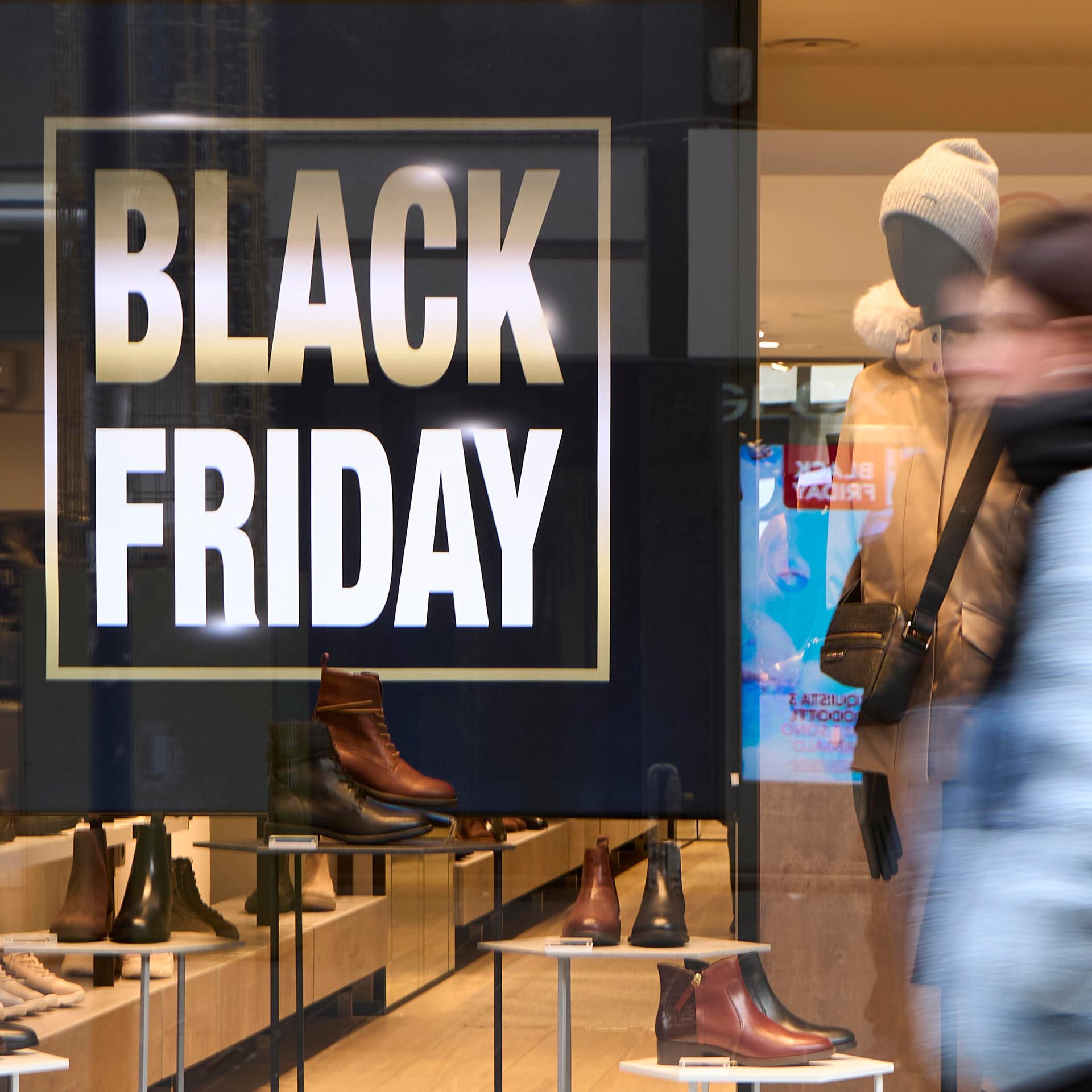 Fußgänger in Winterkleidung laufen an einem hell erleuchteten Schuhladen vorbei; im Schaufenster sind ein großes „BLACK FRIDAY“-Schild und Lederschuhe zu sehen.