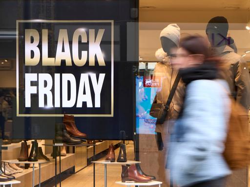 Fußgänger in Winterkleidung laufen an einem hell erleuchteten Schuhladen vorbei; im Schaufenster sind ein großes „BLACK FRIDAY“-Schild und Lederschuhe zu sehen. Fußgänger in Winterkleidung laufen an einem hell erleuchteten Schuhladen vorbei; im Schaufenster sind ein großes „BLACK FRIDAY“-Schild und Lederschuhe zu sehen.