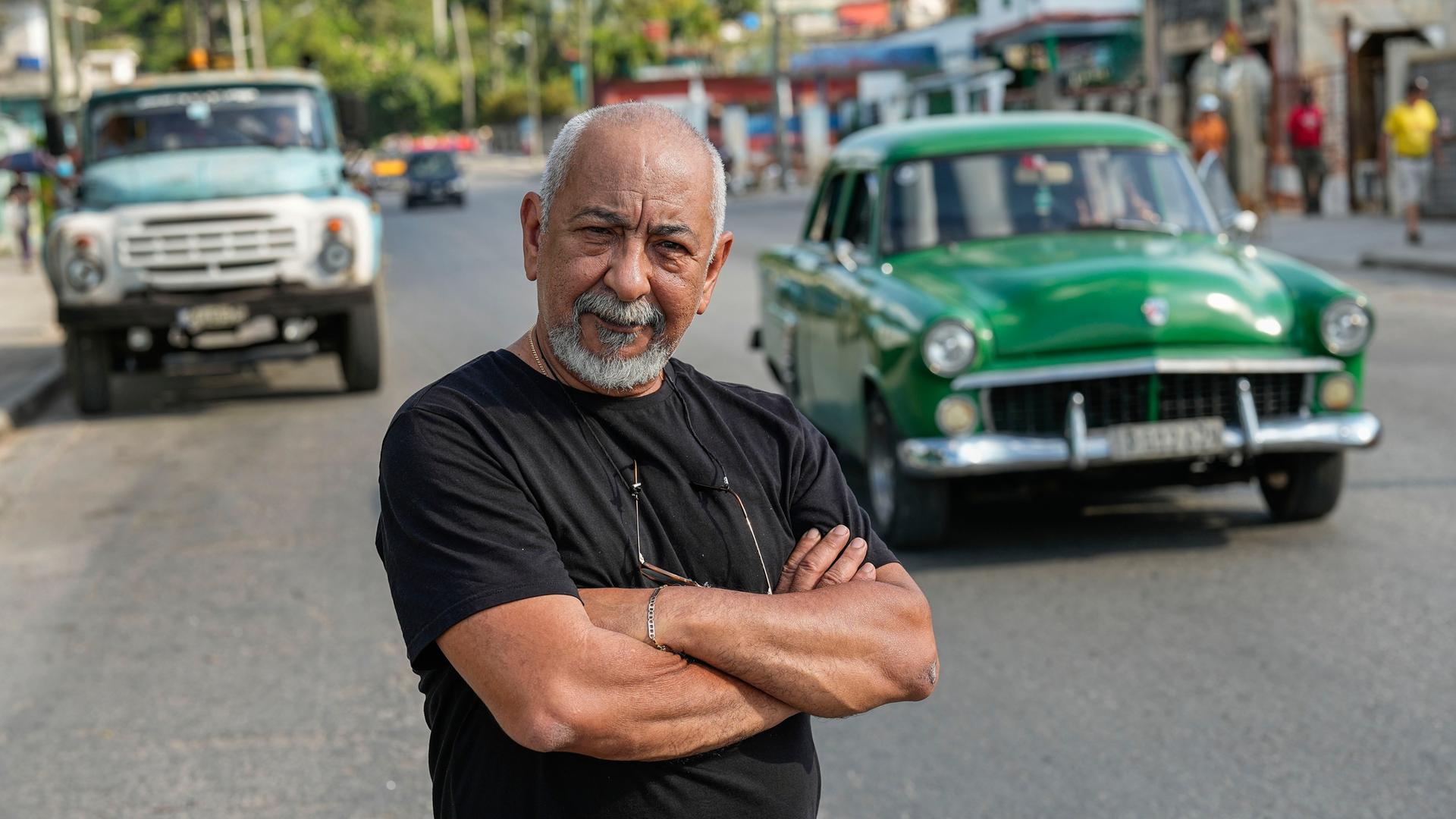 Der kubanische Schriftsteller Leonardo Padura posiert für ein Porträt auf einer Straße in Havanna. Im Hintergrund fährt ein grünes Auto in der für Kuba typischen Anmutung der Fünfzigerjahre.