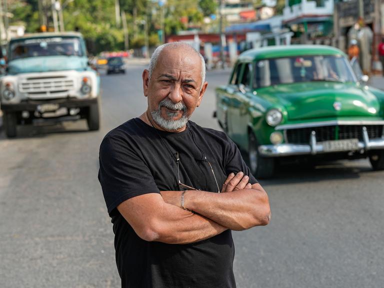 Der kubanische Schriftsteller Leonardo Padura posiert für ein Porträt auf einer Straße in Havanna. Im Hintergrund fährt ein grünes Auto in der für Kuba typischen Anmutung der Fünfzigerjahre.