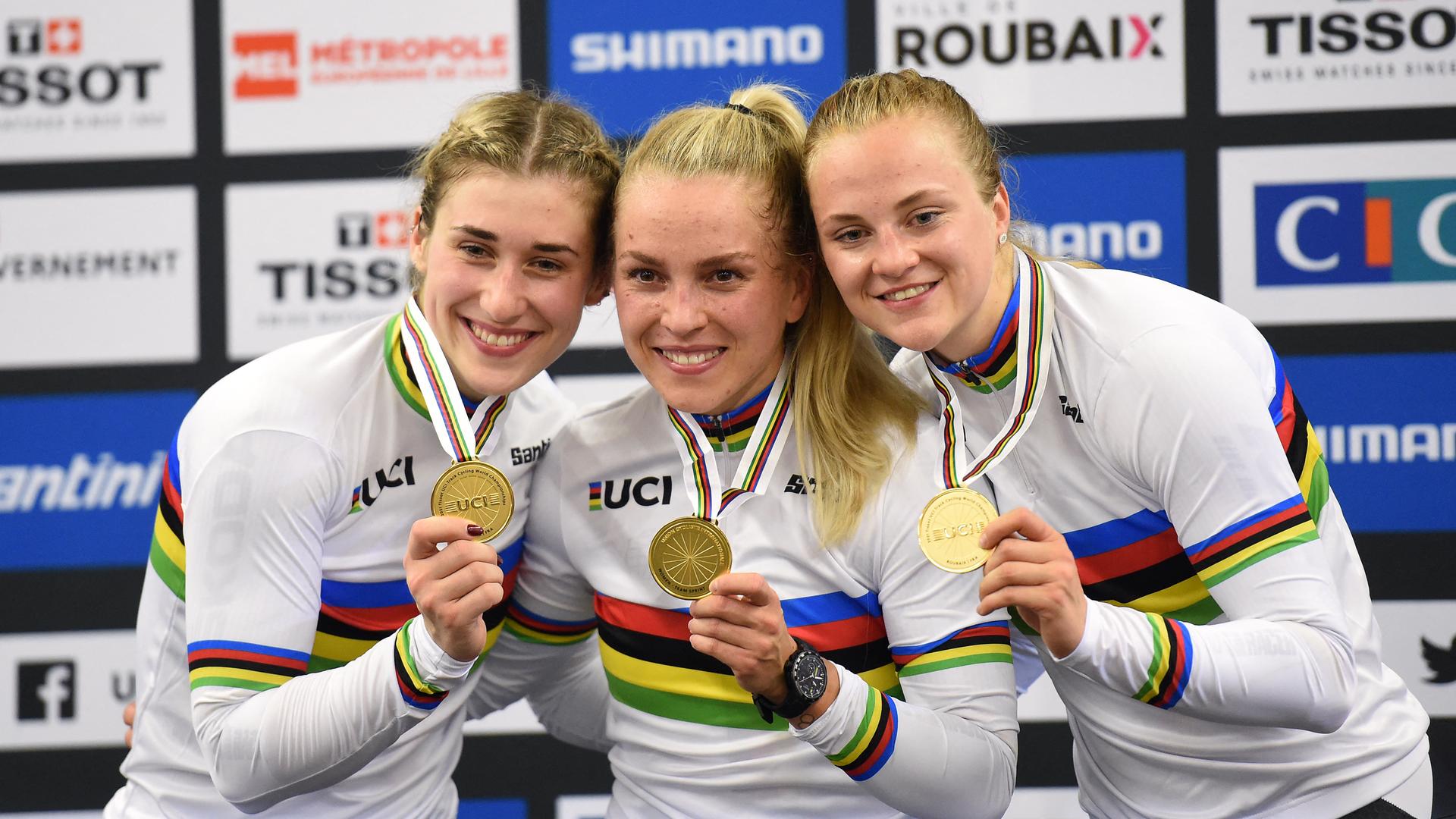 Die drei Sportlerinnen stehen lächelnd eng beieinander und halten ihre Medaillen nach vorne. 