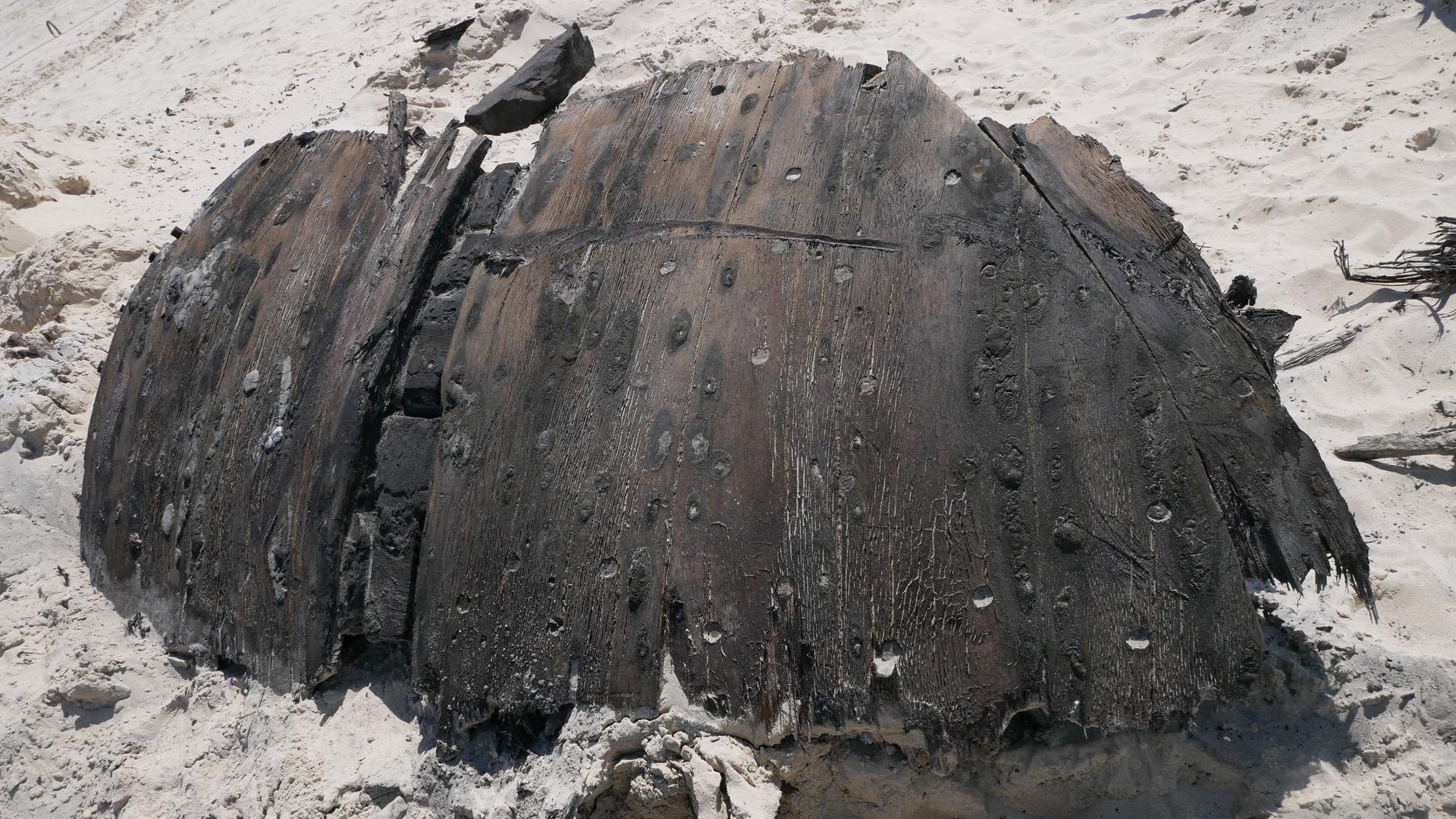 Ein Teil eines rund 300 Jahre alten Schiffswracks liegt am Strand der Nordseeinsel Norderney. Das dunkle Holz ist gebogen. Es liegt im Sand. Ein Teil eines rund 300 Jahre alten Schiffswracks liegt am Strand der Nordseeinsel Norderney. Das dunkle Holz ist gebogen. Es liegt im Sand.