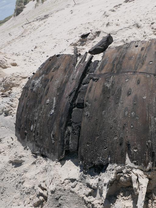 Ein Teil eines rund 300 Jahre alten Schiffswracks liegt am Strand der Nordseeinsel Norderney. Das dunkle Holz ist gebogen. Es liegt im Sand. 