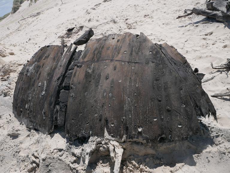 Ein Teil eines rund 300 Jahre alten Schiffswracks liegt am Strand der Nordseeinsel Norderney. Das dunkle Holz ist gebogen. Es liegt im Sand. 