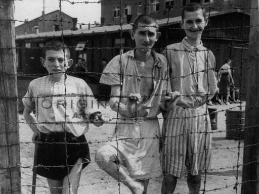 Drei ausgemergelte jugendliche Häftlinge hinter einem Zaun, nach der Befreiung des Konzentrationslagers Buchenwald am 13.04.1945 durch die 3. US-Armee.