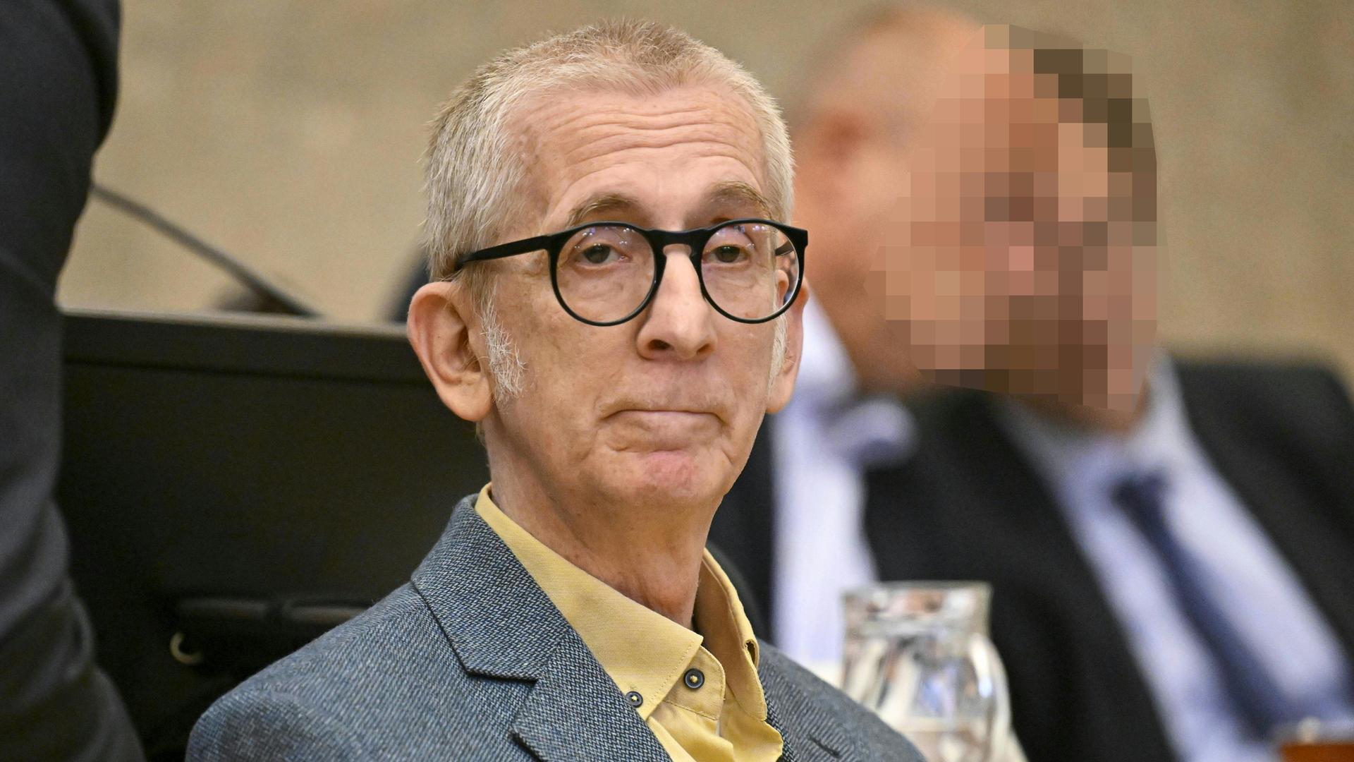 Paal sitzt mit ernstem Blick auf der Anklagebank Er trägt eine runde Brille, kurzes Haaar und ein graues Jackett. Dahinter weitere teils verpixelte Prozessteilnehmer. 
