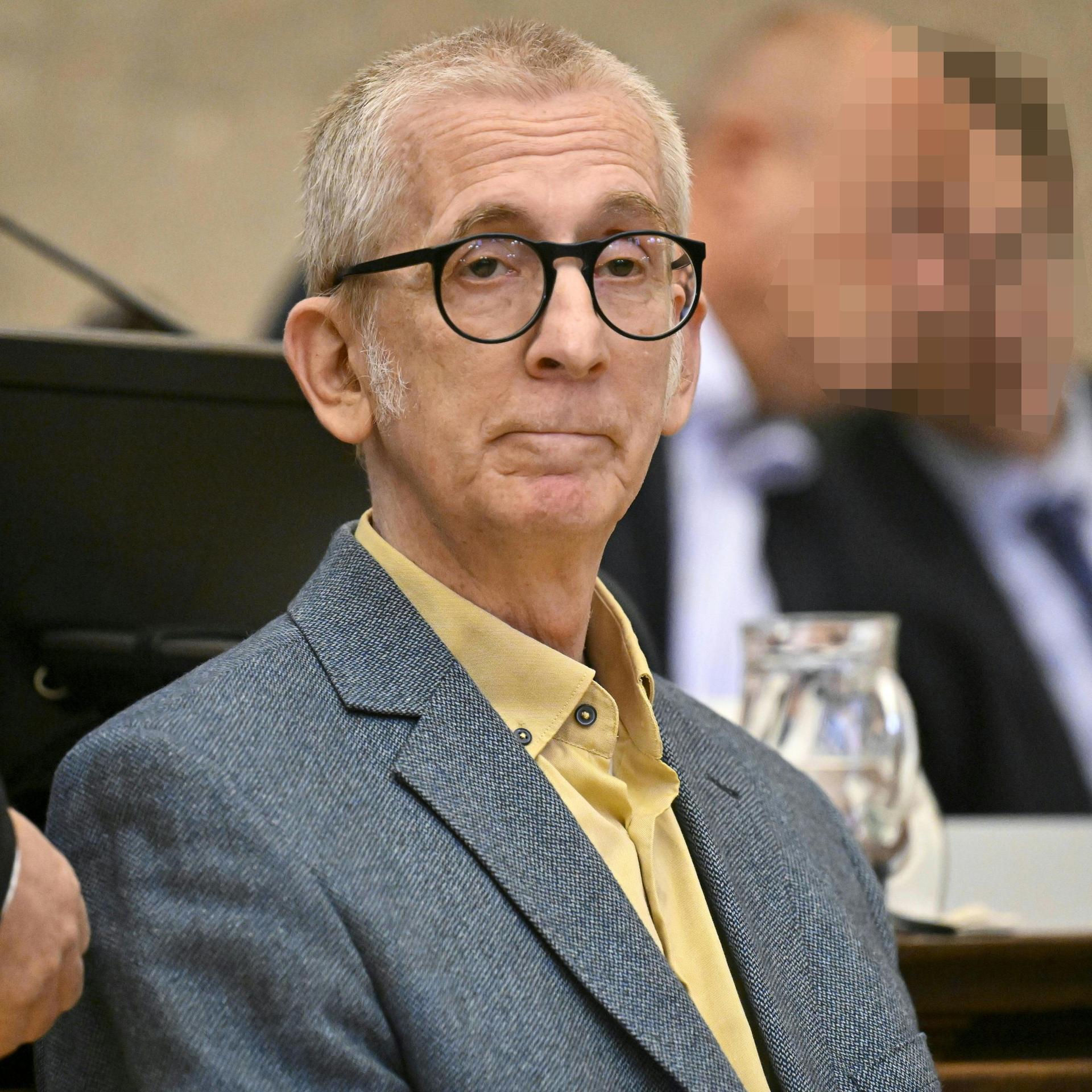 Paal sitzt mit ernstem Blick auf der Anklagebank Er trägt eine runde Brille, kurzes Haaar und ein graues Jackett. Dahinter weitere teils verpixelte Prozessteilnehmer. 