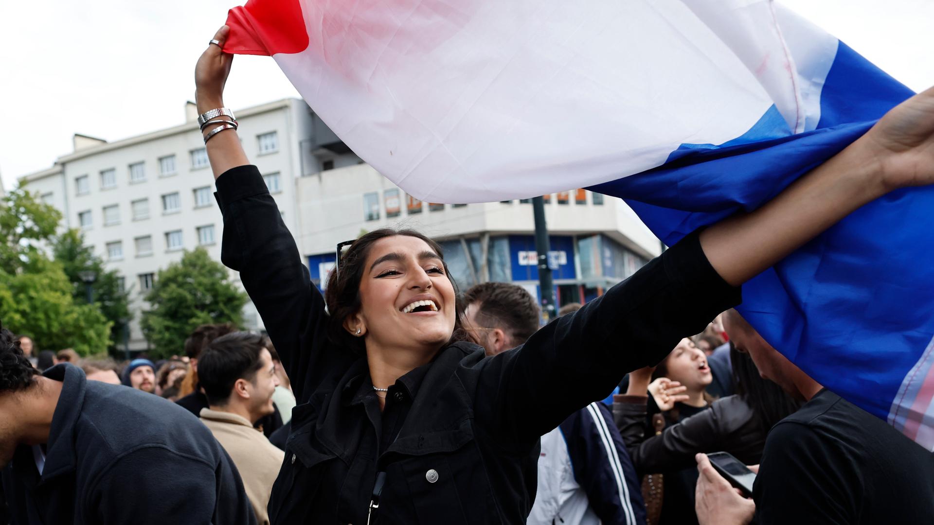 Eine Frau schwenkt die französische Flagge. Die Frau hat dunkle Haare. Sie ist auf einer Demonstration. Die Frau freut sich, dass linke Parteien die Wahl in Frankreich gewonnen haben.