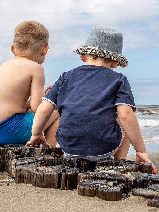 Das Foto zeigt zwei kleine Kinder in Rückenansicht:. Sie  sitzen auf Holzpfählen am Strand. Ein Kind trägt einen Sonnenhut. Strand und Meer sind leer.