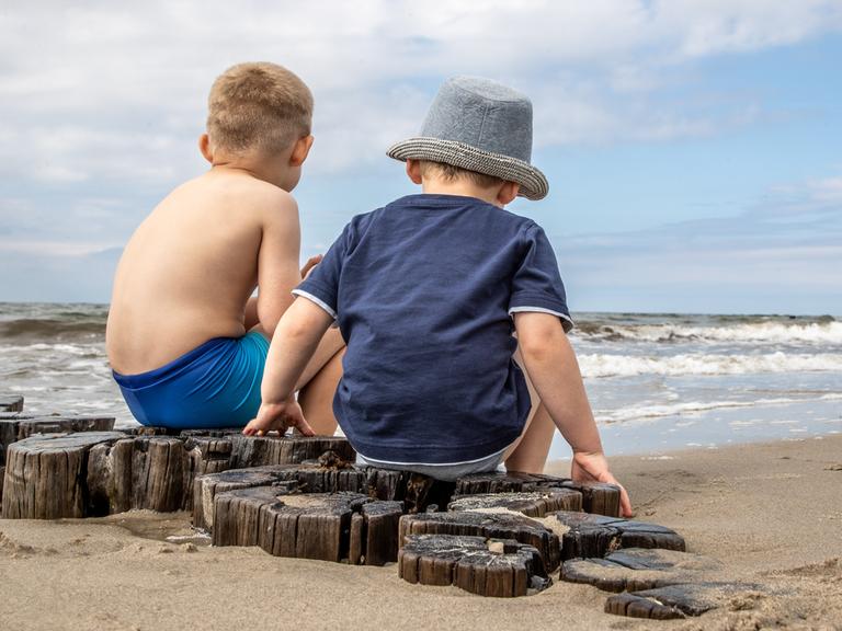 Das Foto zeigt zwei kleine Kinder in Rückenansicht:. Sie  sitzen auf Holzpfählen am Strand. Ein Kind trägt einen Sonnenhut. Strand und Meer sind leer.