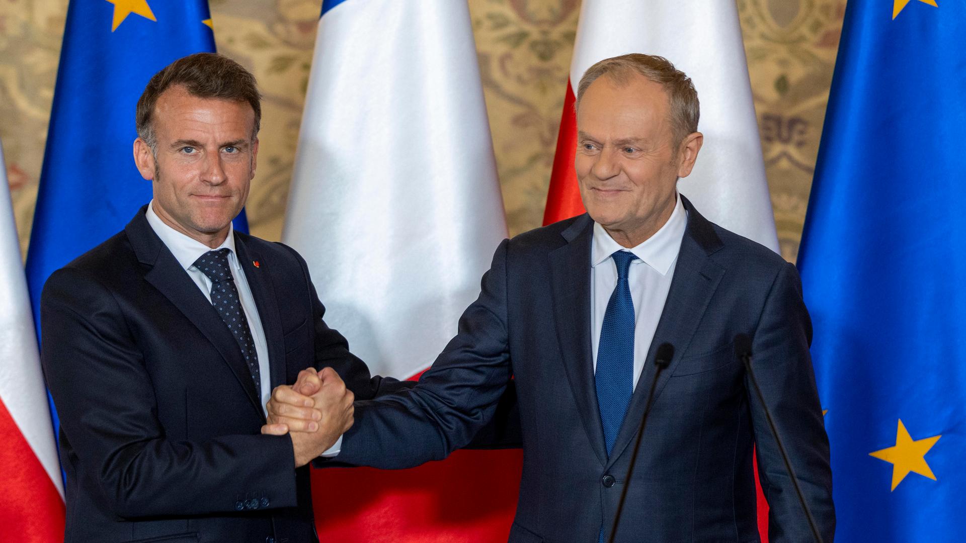 Das Foto zeigt den polnischen Ministerpräsident Tusk (links) und den französischen Präsidenten Macron (rechts), im Hintergrund sind die französische, polnische und die EU-Flagge 