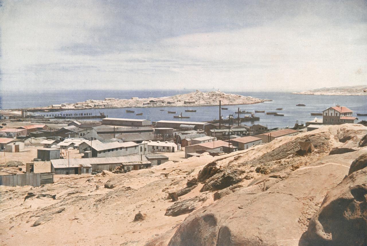 Historische Aufnahme von Lüderitz mit Gebäuden in Küstennähe und der langgestreckten Haifischinsel im Hintergrund.