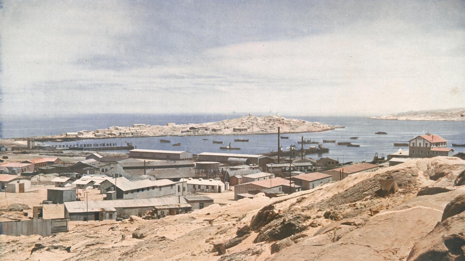Historische Aufnahme von Lüderitz mit Gebäuden in Küstennähe und der langgestreckten Haifischinsel im Hintergrund. Historische Aufnahme von Lüderitz mit Gebäuden in Küstennähe und der langgestreckten Haifischinsel im Hintergrund.