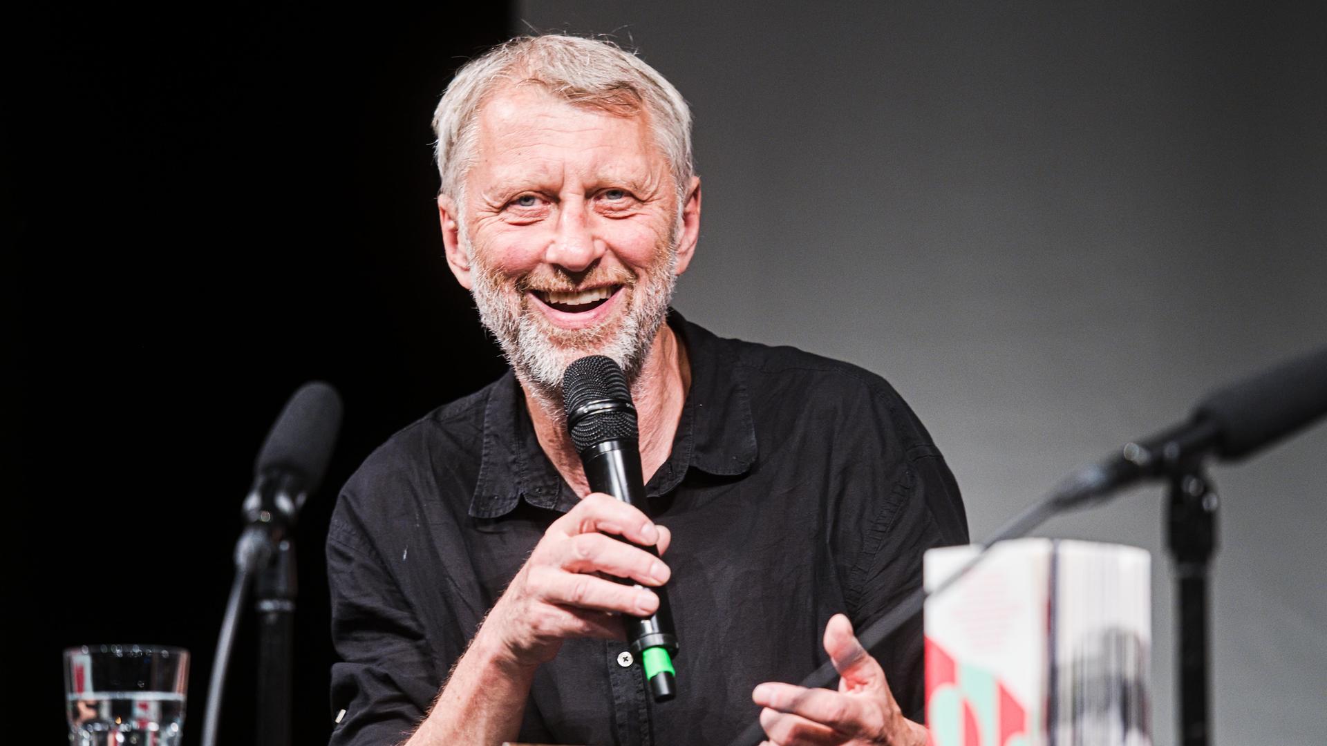 Jáchym Topol bei einer Lesung. Er lächelt freundlich in die Kamera. Er hat graue Haare und ein Mikrofon in der Hand. Jáchym Topol bei einer Lesung. Er lächelt freundlich in die Kamera. Er hat graue Haare und ein Mikrofon in der Hand.