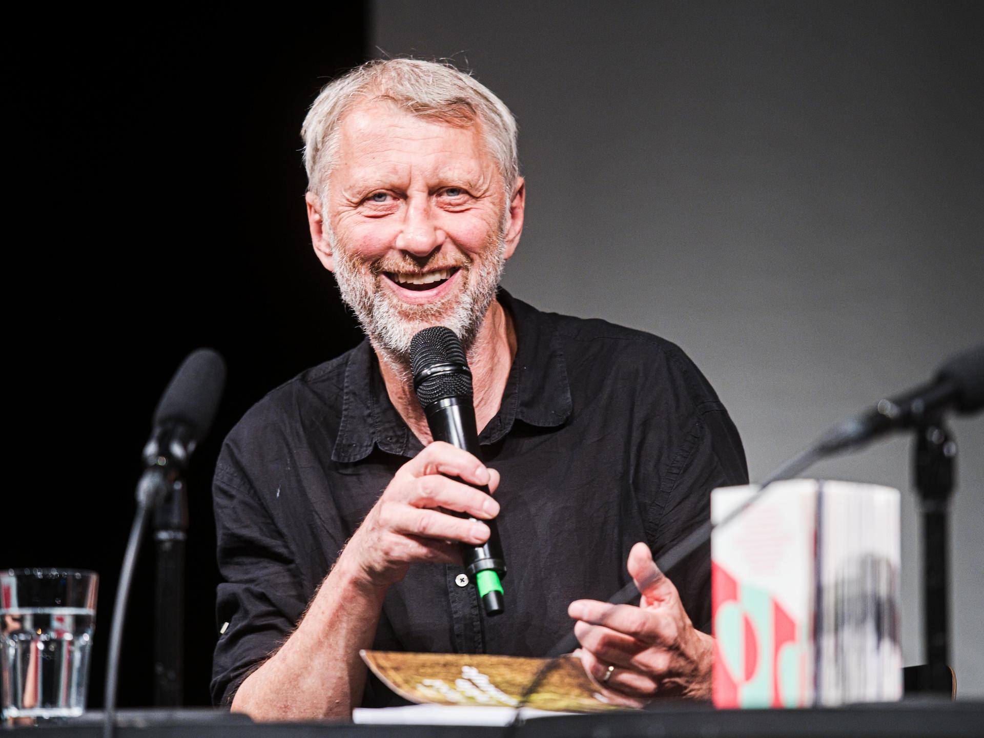 Jáchym Topol bei einer Lesung. Er lächelt freundlich in die Kamera. Er hat graue Haare und ein Mikrofon in der Hand. 
