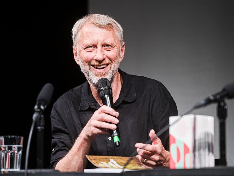 Jáchym Topol bei einer Lesung. Er lächelt freundlich in die Kamera. Er hat graue Haare und ein Mikrofon in der Hand. 
