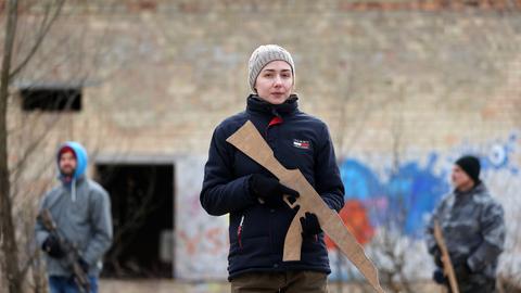 Eine junge Frau, mit heller Mütze und einer dunklen Jacke, steht vor einem alten Backsteingebäude. In ihren Händen hält sie eine Waffenattrappe und schaut den Betrachter direkt an.