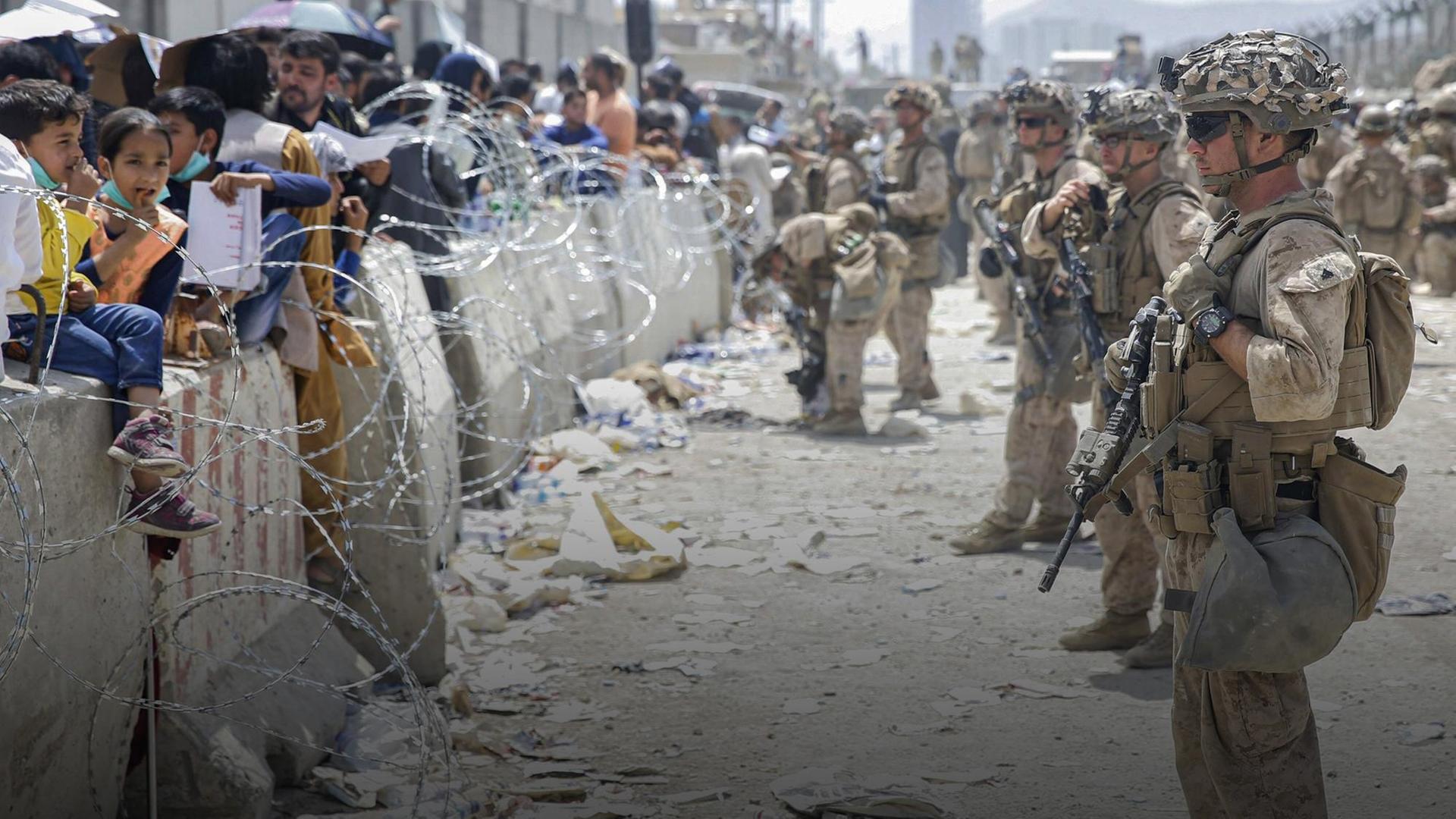 Bewaffnete Soldaten sichern den Flughafen Kabul in Afghanistan 2021, vor den Mauern stehen zahlreiche Menschen und Kinder hinter Stacheldraht.
