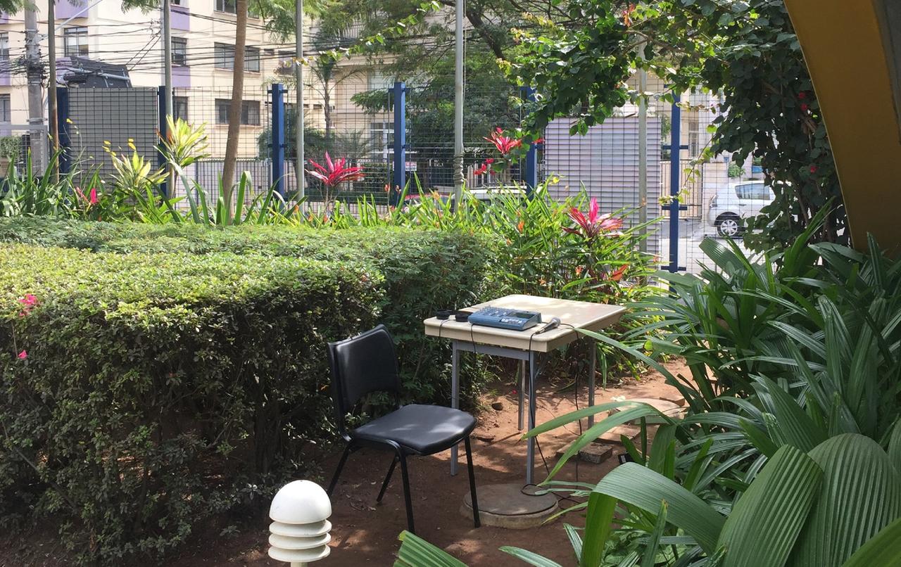 In einem städtischen Garten, etwas abgeschottet unter Bäumen und neben Hecken, im Hintergrund ein Zaun: Ein Stuhl und ein Tisch mit Recorder, Mikrofon und Kopfhörern. 