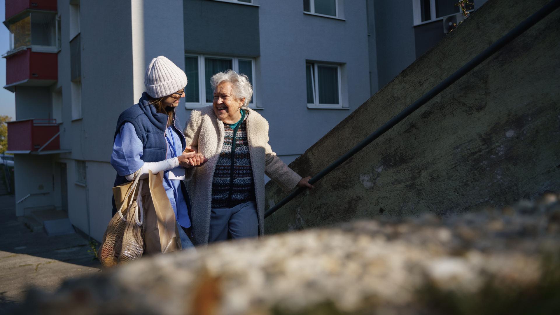 Eine Frau begleitet eine Seniorin am Arm die Stufen vor einem Wohnhaus hinauf. Auf dem Arm trägt sie Papiertaschen mit Einkäufen.
Eine Frau begleitet eine Seniorin am Arm die Stufen vor einem Wohnhaus hinauf. Auf dem Arm trägt sie Papiertaschen mit Einkäufen.